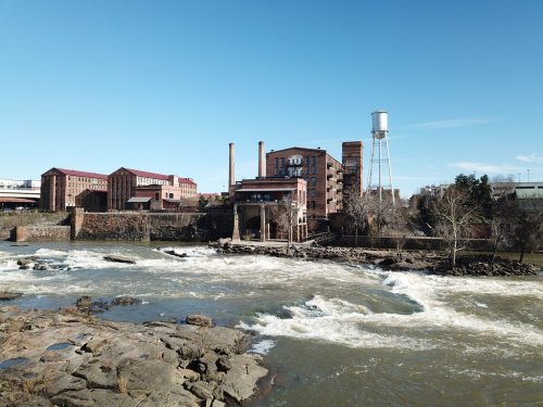 A river flows past an old industrial building and water tower under a clear blue sky.