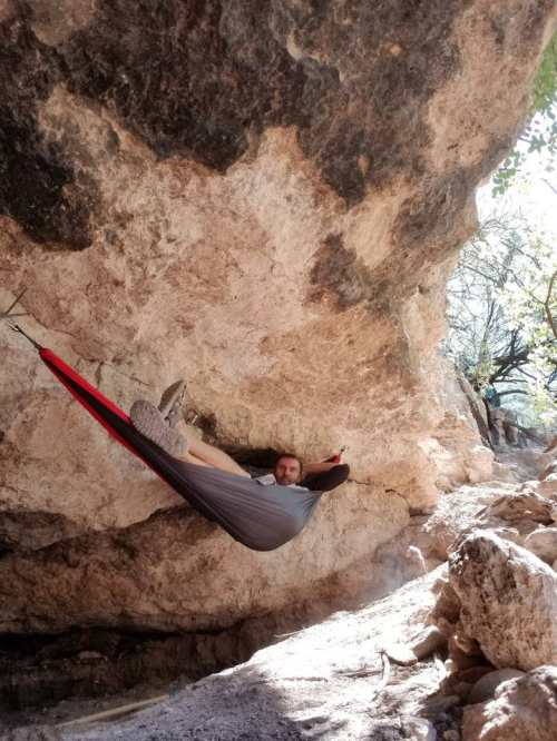 A person relaxes in a hammock under a rocky overhang in a natural outdoor setting.