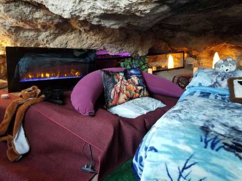 Cozy cave interior with a bed, cushions, and decorative lighting, featuring a faux fireplace and soft blankets.