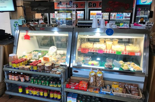 A seafood market display with fresh fish, salads, and snacks in glass cases, surrounded by various condiments and drinks.