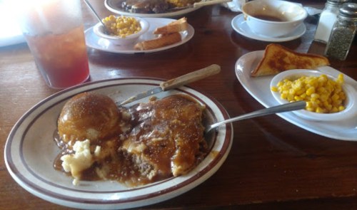A plate of meatloaf with gravy, mashed potatoes, and a roll, accompanied by corn and toast on a wooden table.