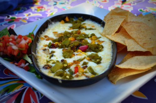 A black dish of melted cheese topped with green chiles, served with tortilla chips and a side of fresh salsa.