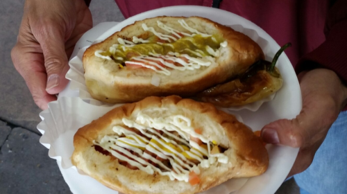A person holds a plate with two hot dogs topped with mustard, ketchup, mayonnaise, and sliced vegetables, alongside a pepper.