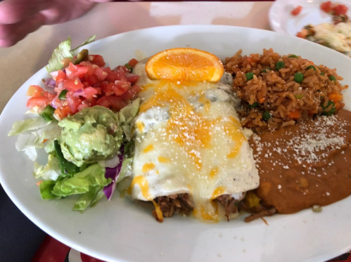 A plate of enchiladas topped with cheese, served with rice, beans, salad, guacamole, and a slice of orange.
