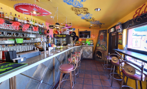 Colorful bar interior with a long counter, stools, and vibrant decorations, including sombreros and artwork on the walls.