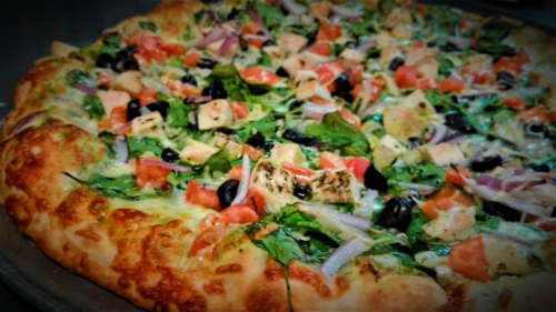 A close-up of a colorful pizza topped with greens, tomatoes, onions, olives, and chunks of chicken on a golden crust.