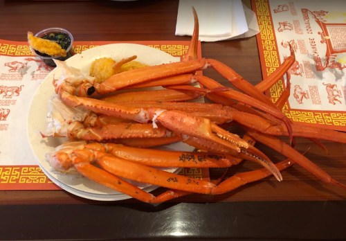 A plate of large, orange crab legs with a side of dipping sauce and a fried item, set on a patterned tablecloth.