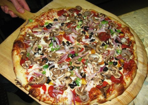A freshly baked pizza topped with pepperoni, mushrooms, onions, green peppers, and black olives on a wooden pizza peel.