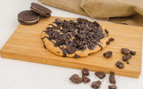 A large chocolate chip cookie topped with crushed Oreos and drizzled with chocolate, placed on a wooden cutting board.