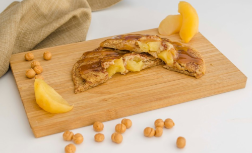 A wooden board with a sliced pastry filled with apples, drizzled with caramel, and scattered hazelnuts.