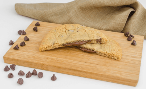 A chocolate chip cookie split in half, revealing a gooey chocolate filling, on a wooden cutting board with chocolate chips scattered.