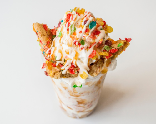 A tall dessert glass filled with ice cream, topped with whipped cream, colorful cereal, and a cookie.