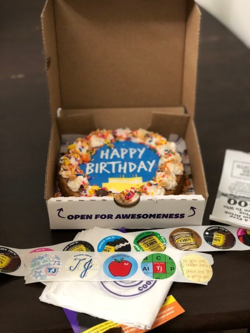 A birthday cake in a box with colorful sprinkles and a "Happy Birthday" sign, surrounded by fun stickers and napkins.