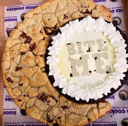 A large cookie with chocolate chips surrounds a cake that says "BITE ME" in the center, topped with whipped cream.