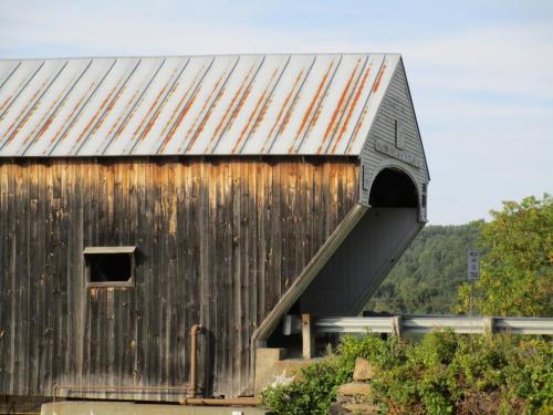 A weathered covered bridge with a metal roof, partially obscured by greenery and set against a blue sky.