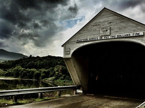 A covered bridge with a sign reading "Walk your horses or pay two dollars fine," set against a cloudy sky and lush landscape.