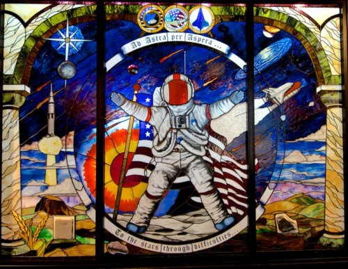 Stained glass mural featuring an astronaut with flags, rockets, and celestial imagery, symbolizing exploration and perseverance.