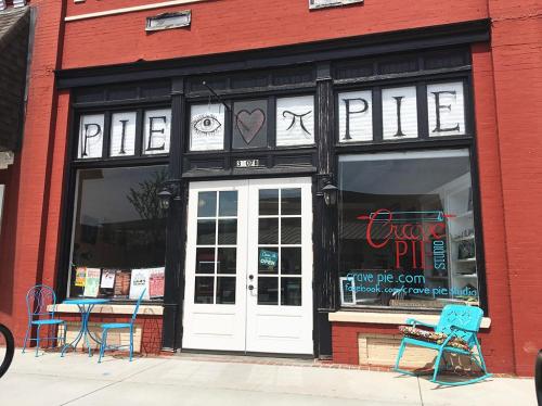 A storefront with large windows displaying "PIE ♥ PIE" and a sign for Crave Pie Studio, featuring outdoor seating.
