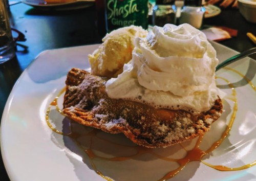 A dessert featuring a fried pastry topped with whipped cream and a scoop of ice cream, drizzled with caramel sauce.