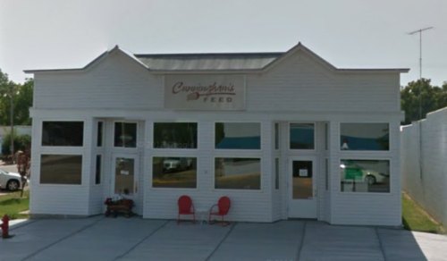 A white, two-story building with large windows and a sign reading "Cunningham's Feed," featuring red chairs outside.