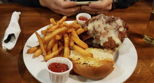 A plate with a sandwich topped with fried chicken and cheese, served with fries and ketchup on a wooden table.