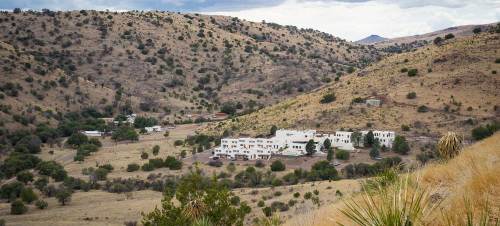 A white building nestled in a hilly, dry landscape with sparse vegetation and distant mountains under a cloudy sky.