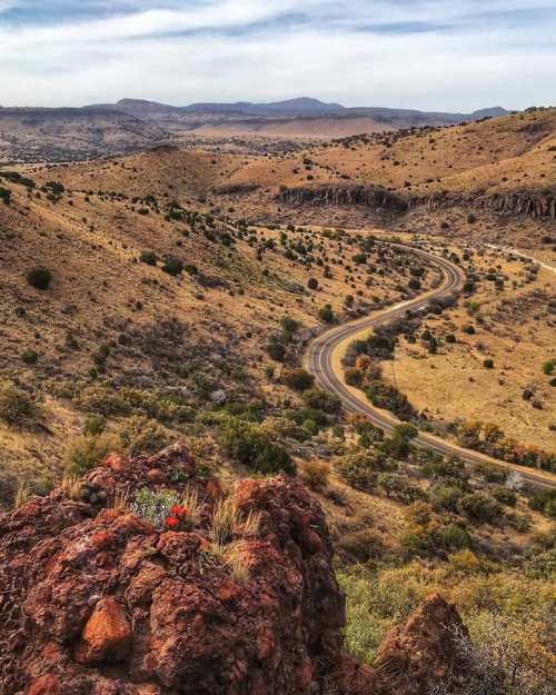 A winding road cuts through a dry, hilly landscape with sparse vegetation and distant mountains under a blue sky.