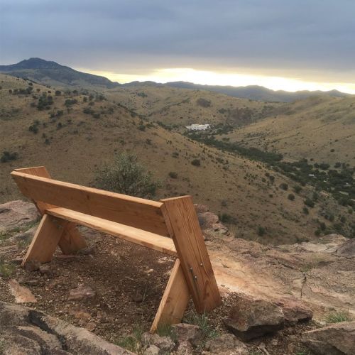A wooden bench overlooks a mountainous landscape under a cloudy sky at sunset.