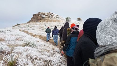 A group of people walking on a frosty trail towards a rocky hill, surrounded by white grass and a cloudy sky.