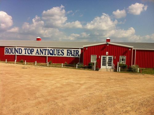 Red building with a sign reading "Round Top Antiques Fair," set against a cloudy sky and gravel parking area.