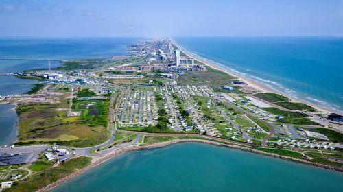 Aerial view of a coastal area with a long beach, city skyline, and green spaces along the shore.