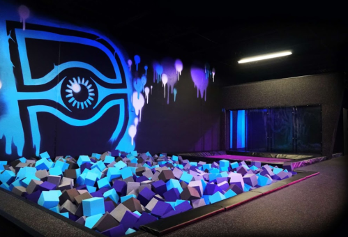 A colorful indoor trampoline park featuring a foam pit with blue and gray blocks and vibrant wall art.