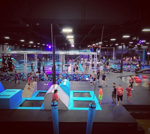 A vibrant indoor trampoline park filled with people enjoying various activities and obstacles under colorful lights.