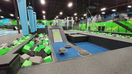 Indoor trampoline park featuring foam pits, climbing walls, and various jumping areas with vibrant green and blue decor.