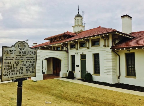 Historic sign in front of a white building with a red-tiled roof, marking the site of the first railroad in the area.