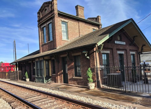 Historic brick train station with a green roof, surrounded by railroad tracks and potted plants under a clear blue sky.