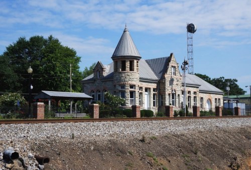 Historic stone train station with a turret, surrounded by greenery and a gravel track under a blue sky.