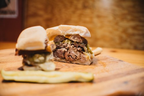 A close-up of a sandwich with meat, cheese, and toppings, served on a wooden board with a pickle on the side.