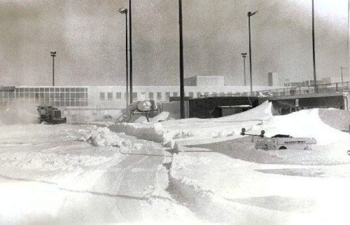 A snowy landscape with large snowdrifts, machinery clearing paths, and a building in the background under a gray sky.