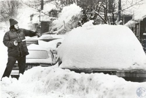 A person shovels snow off a car in a snowy street, surrounded by deep snow and parked vehicles.