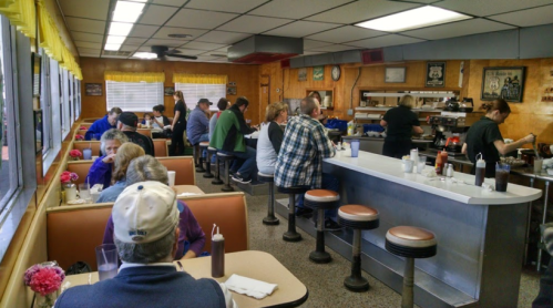 A busy diner interior with patrons seated at booths and the counter, enjoying meals and drinks in a cozy atmosphere.