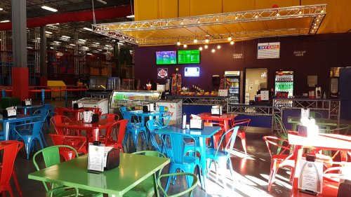 Brightly colored tables and chairs in a cafe area with a counter, screens, and vending machines in a recreational facility.