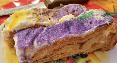 A colorful king cake with purple, yellow, and green icing, sliced and served on a decorative plate.