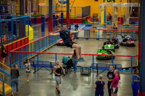A man swings joyfully above a colorful indoor play area filled with people enjoying various activities.