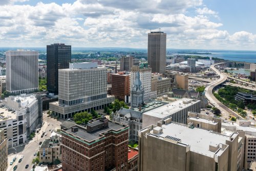 Aerial view of a city skyline with modern buildings, a highway, and a lake in the background under a partly cloudy sky.