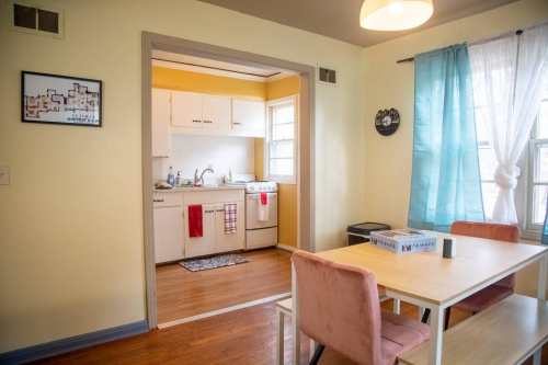 A cozy dining area with a wooden table and pink chairs, leading to a bright kitchen with white cabinets.