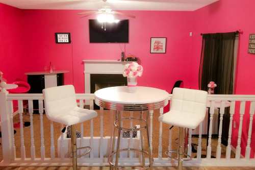 A bright pink room featuring a round table with two white stools, a TV on the wall, and decorative flowers.