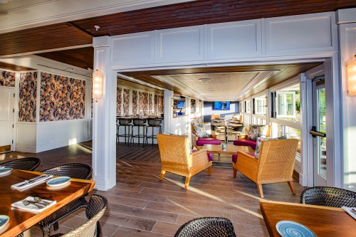 Brightly lit restaurant interior featuring wicker chairs, wooden tables, and tropical wallpaper. Bar area visible in the background.