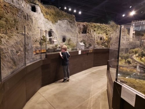 A child observes a detailed model train display in a museum, surrounded by scenic miniature landscapes.