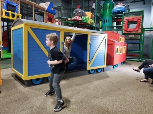 A child plays near a colorful train structure while an adult poses playfully from inside the train.